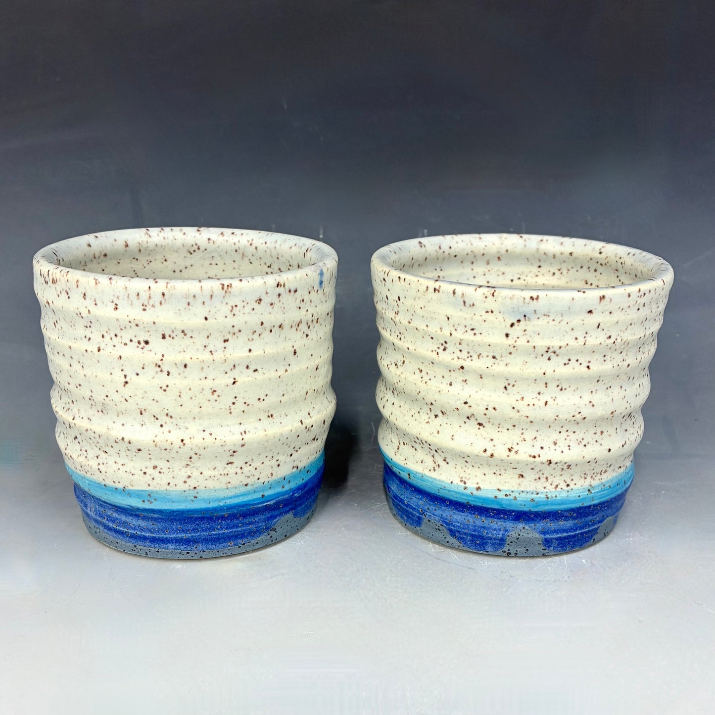 Two ceramic cups with textured white bodies and blue bands on a gray background