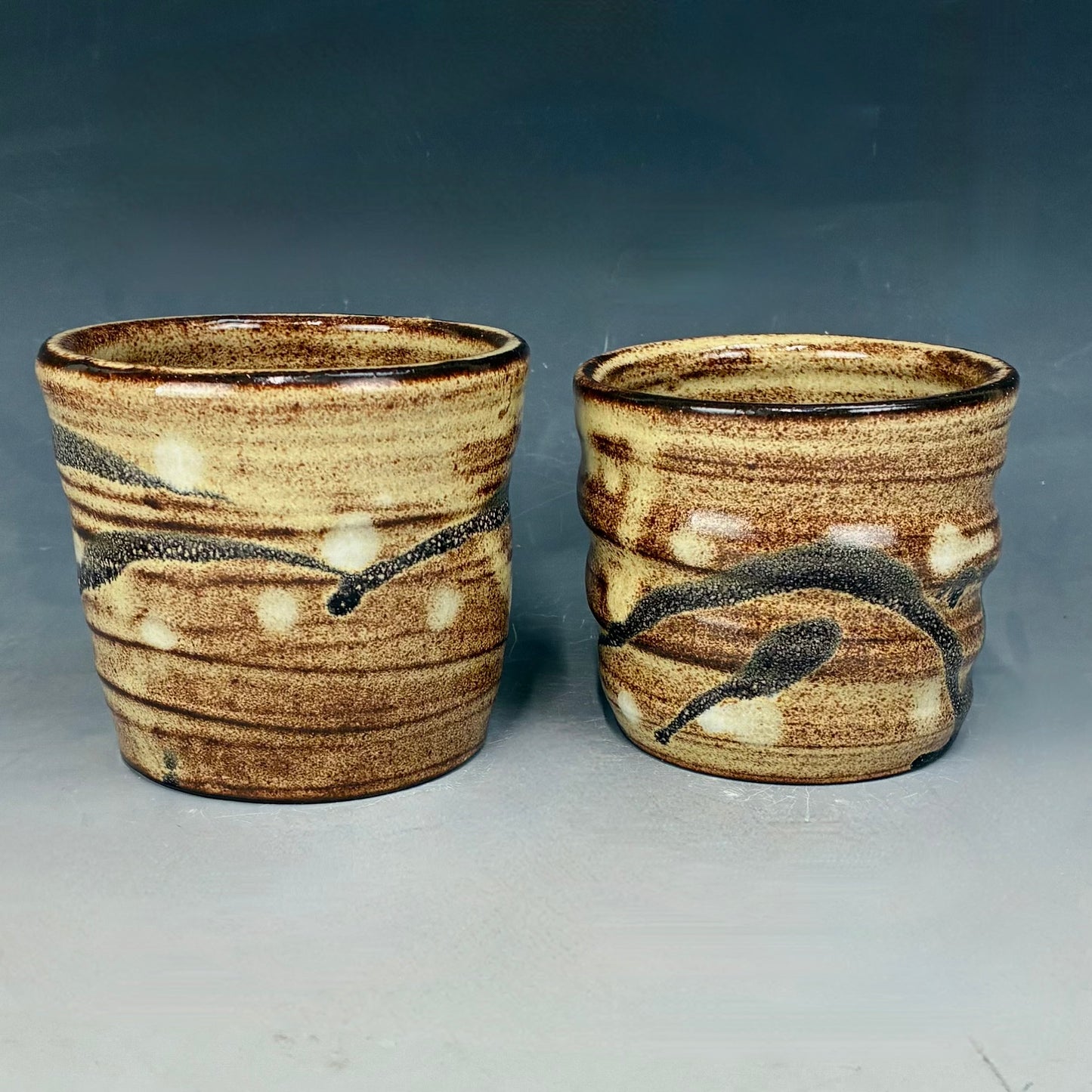 Two ceramic cups with brown and beige glaze on a gray background